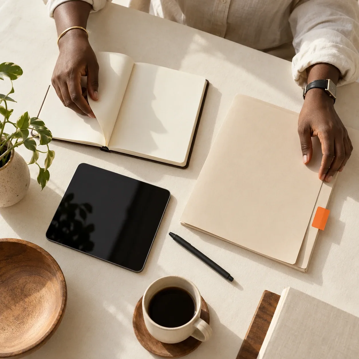 Black hands organising a notebook, folder and tablet on a cream desk.